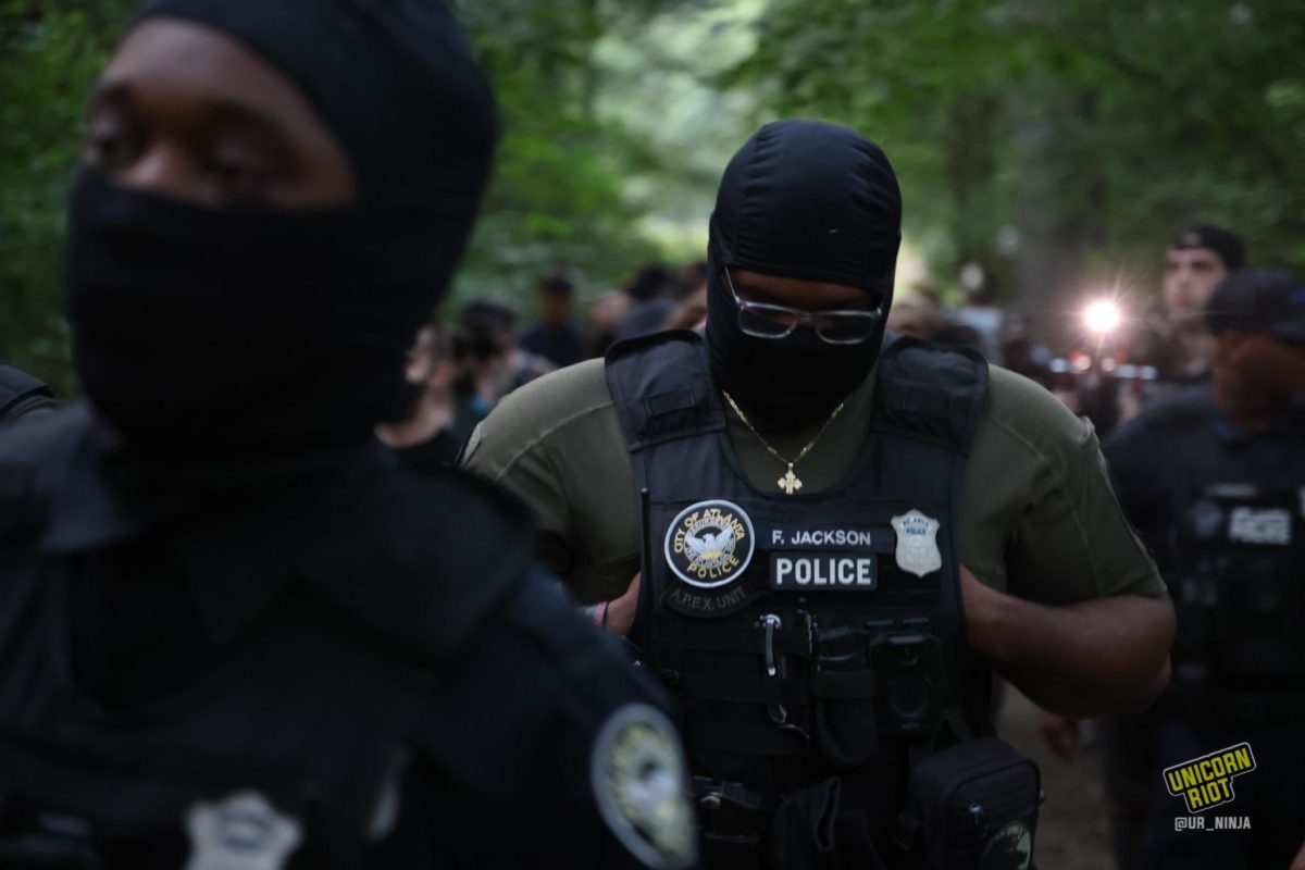 Atlanta Police officers in tactical vests and balaclava-like face masks sweep through the park as the vigil for Manuel ‘Tortuguita’ Terán was set to begin at 8:30 p.m. One officer holds his head facing down with with the frames of his eyeglasses visible in the eye cutout of his mask. His vest features the nametag 'F. Jackson' and to its left is a patch with the seal of the Atlanta Police Department. Below the APD logo patch is a smaller patch reading "A.P.E.X. Unit in very faint green-grey lettering.