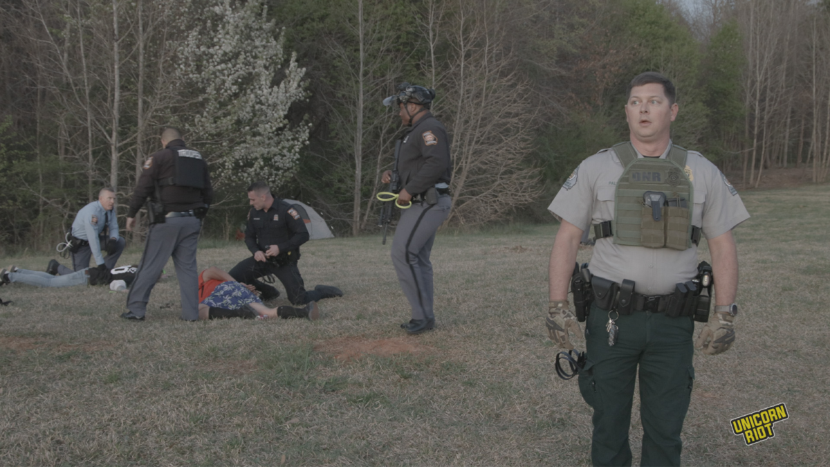 A white Georgia Department of Natural Resources game warden wears a small bulletproof vest with his belly protruding underneath looks to the side with his mouth agape, his face featuring a slightly phased and aloof look. Behind him and to the viewer's left, two people are face down in the process of being arrested by 4 Goergia state troopers - three troopers are white, one is black. They are wearing brown, black or light blue uniform shirts with pin striped gray pants. Most of them have plastic zip tie 'flex-cuffs' in hand as part of the arrest process taking place in the RC Field area of the South River Forest during the 'Stop Cop City' music festival on March 5, 2023. Trees can be seen in the forest behind the arrest scene; all is bathed in a pale faintly orange light as the sun begins to set. 