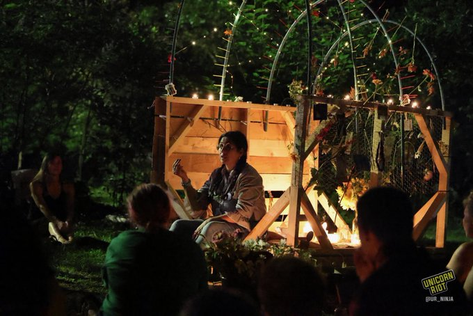Belkis Terán addresses the crowd at the nighttime vigil at Brownwood Park while seated on a dimly lit wooden structure with metal hoops with plants and flowers hanging above where she is sitting.
