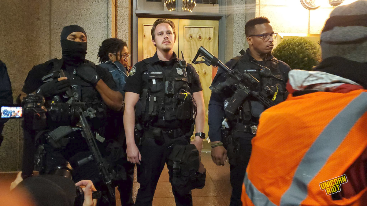 Heavily armed Atlanta Police officers guard the headquarters of the Atlanta Police Foundation at 191 Peachtree Street during a Black-led protest on Mach 9, 2023. A white male officer with nametag 'A. Luebbehusen' looks at the crowd looks at the crowd with what appears to be disdain or disgust on his face. He is flanked by two Black male officers - one masked, one unmasked with eyeglasses - both of whom have assault rifles strapped to their chests. 