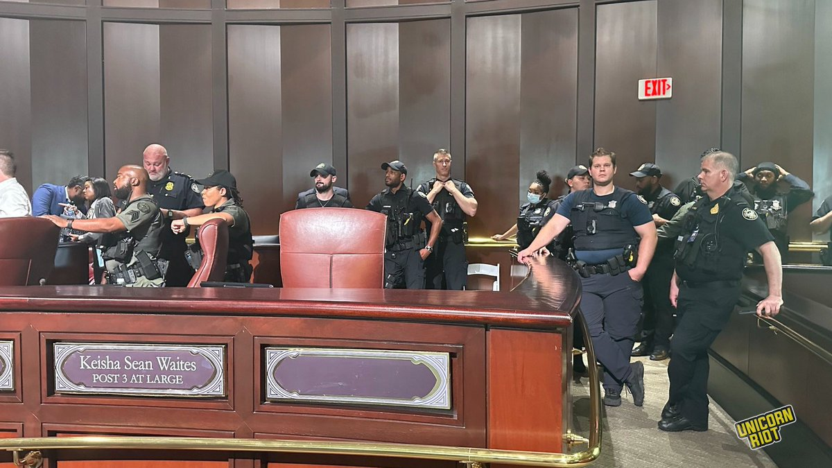 13 Atlanta Police officers stand behind the empty seat in Atlanta City Council chambers labeled" Keisha Sean Waites Post 3 At Large. Sgt. Neil Welch leans against a waist-high wall gazing into the distance with sweat on his face and his cell phone in his right hand. To his left a younger officer with a lot of hair gel in his hair stares menacingly directly into the camera.