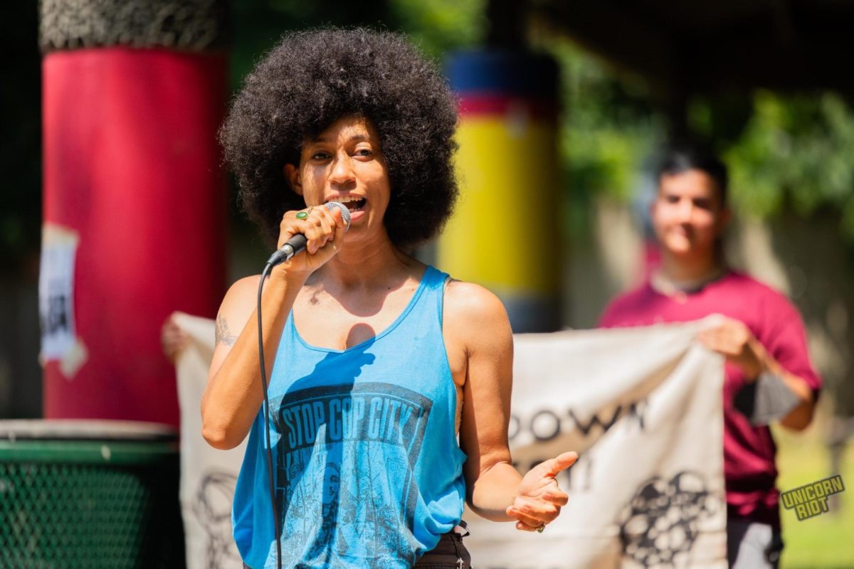 Dr. Mariah Parker (they/them), an African-American femme-presenting person wearing a blue ‘Stop Cop City’ tank top with an Afro haircut speaks into a wired microphone standing in front of other protesters holding an out-of-focus banner held from white cloth in front of the shaded gazebo/picnic table area at Brownwood Park in southeast Atlanta. The sun is coming down brightly as it’s around noon.