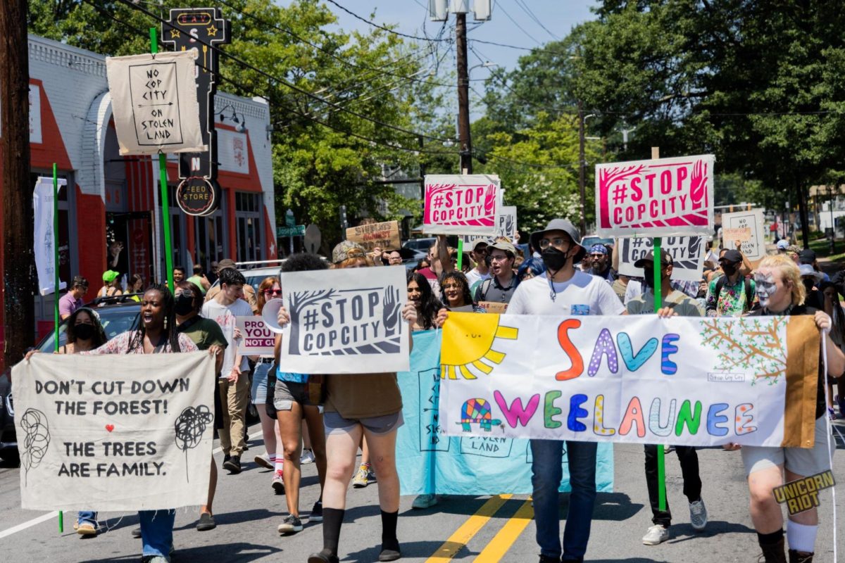 A crowd of about 100 seen from the front marching in the street in the East Atlanta Village neighborhood. Several signs read “Stop Cop City” - two banners at the front of the March read “Don’t Cut Down the Forest! The Trees Are Family.” And “Save Weelaunee” alongside children’s drawings of the sun, a tree and a turtle.