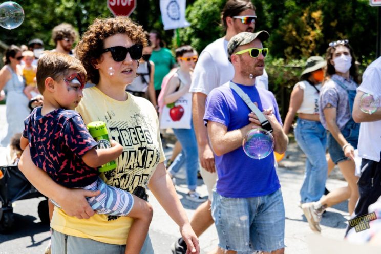 A white child with Spider-Man-style face paint over their eyes is held by their parent as they walk amidst the larger crowd of protesters.