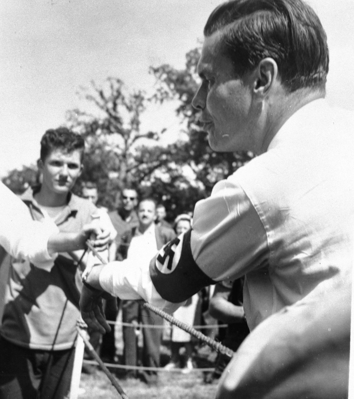 American Nazi Party leader George Lincoln Rockwell shown in a black and white photo addressing a small crowd wearing a swastika armband over the left arm of his white shirt. Rockwell's hair can be seen with a part combed into the left side of his head. Some trees are faintly visible out-of-focus behind the dozen or so spectators seen in frame. 