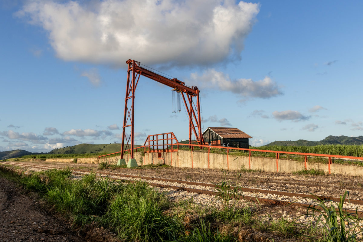 A red crane next to a wooden storehouse set against a cloudy blue sky. A ramp leads up to the crane. A sugar cane field and green mountains lie behind the storehouse.