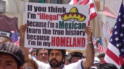 A LAtino man in a white shirt sports medium-short hair and a trimmed full beard while holding a sign that says "If you think I'm "illegal" because I'm a Mexican learn the true history because I'm in my HOMELAND" - the sign also features a white and red sombrero with a pair of eyes peeking out from under it. The man holds the sign amidst a dense crowd of immigrant rights protesters on May Day 2006 in downtown Los Angeles. Photo credit: Jonathan McIntosh, Creative Commons 2.5